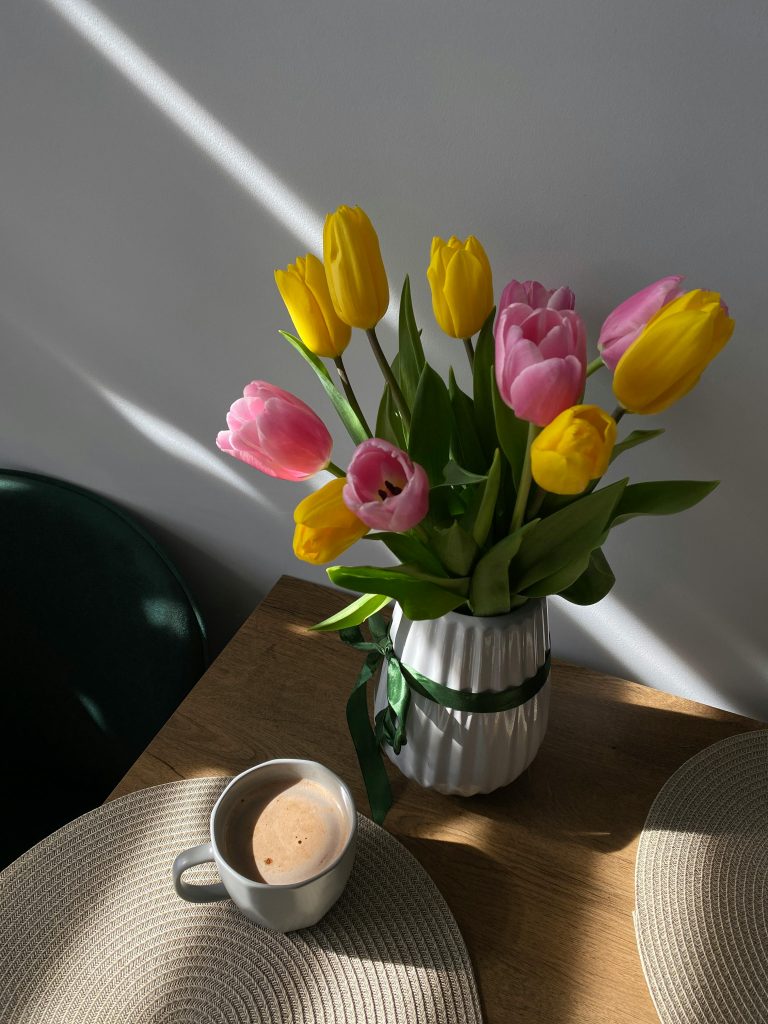 A sunlit arrangement of yellow and pink tulips in a vase with a cup of coffee on a table.